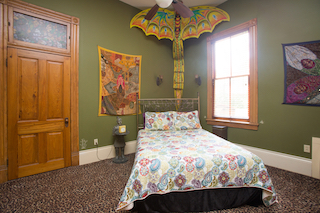 A cozy guest room with a single bed, wooden door, window, and colorful artwork.